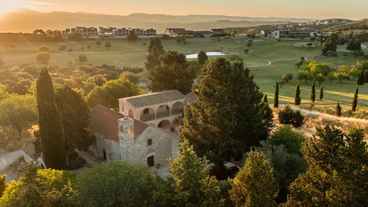 Luftaufnahme eines von Bäumen umgebenen Steingebäudes inmitten einer grünen Landschaft mit Golfplatz – eine ideale Kulisse für eine Golfrundreise durch Irland – sowie moderne Gebäude in der Ferne bei Sonnenuntergang.