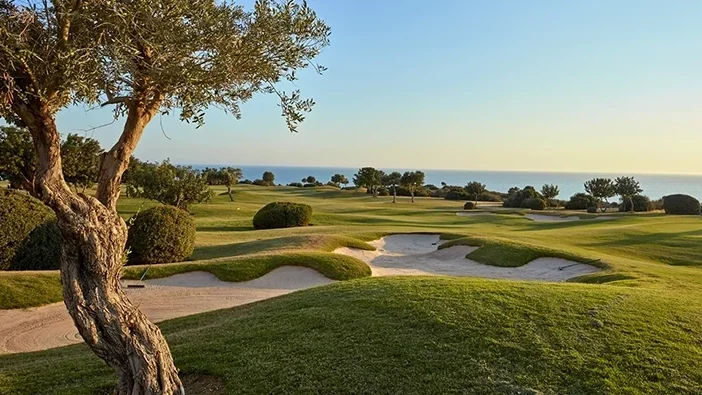 Ein malerischer Golfplatz mit Sandbunkern, saftig grünem Gras und Bäumen, mit Blick auf das Meer unter strahlend blauem Himmel bei Sonnenuntergang – perfekt für ein Abenteuer auf einer Golfrundreise durch Zypern.