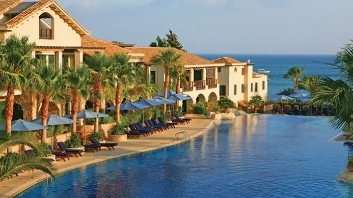 Ein luxuriöses Resort mit Palmen, Liegestühlen und blauen Sonnenschirmen, die einen großen Pool säumen, mit Blick auf das Meer und Boote in der Ferne unter strahlend blauem Himmel – ideal zum Entspannen nach Ihrer Golfrundreise durch Irland.